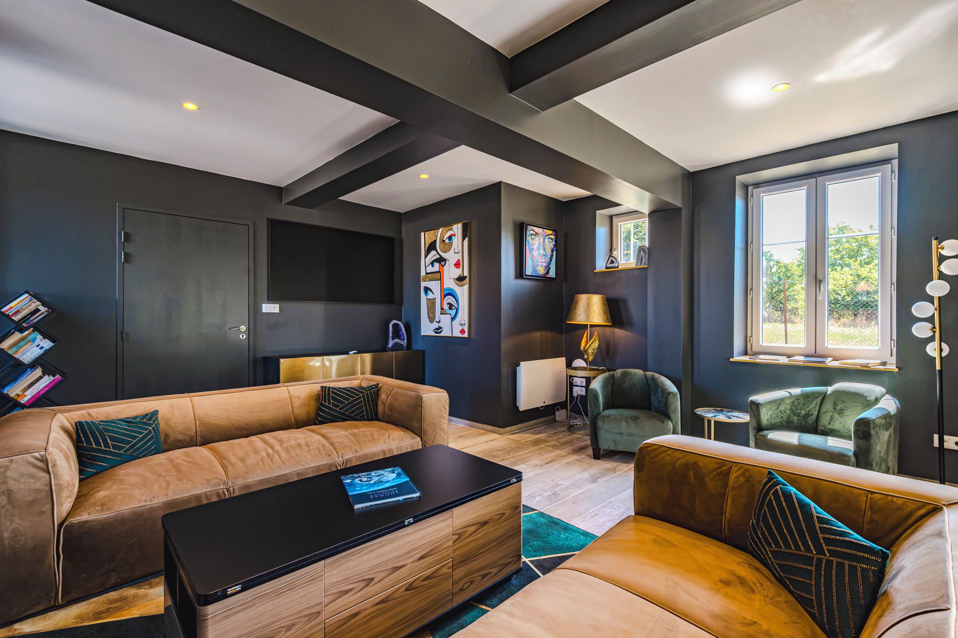 A modern living room with a sofa, coffee table, and armchairs, featuring artwork and a large window.