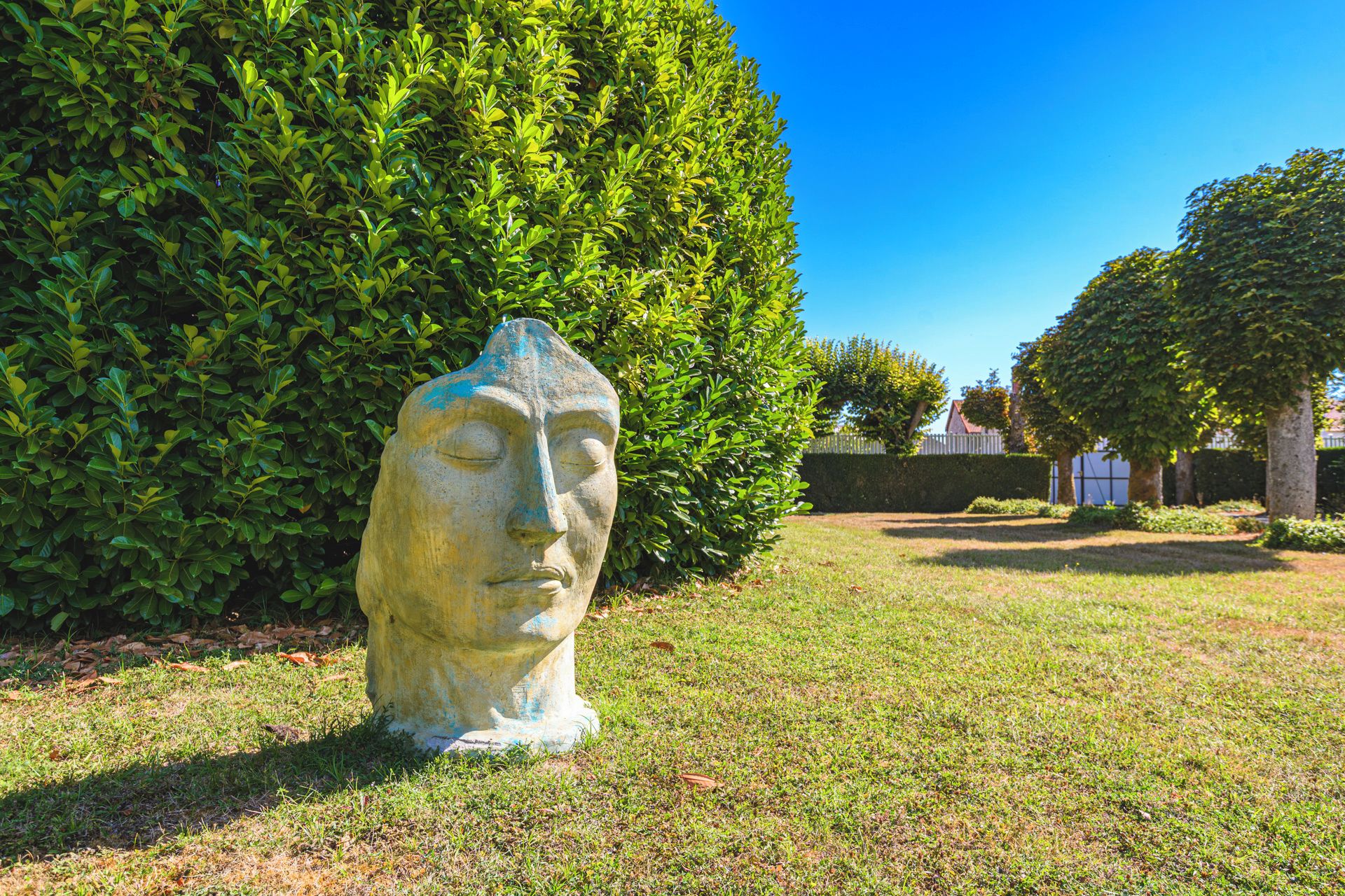 Eine Steinskulptur in einer grünen Umgebung mit einer Hecke und Bäumen im Hintergrund.