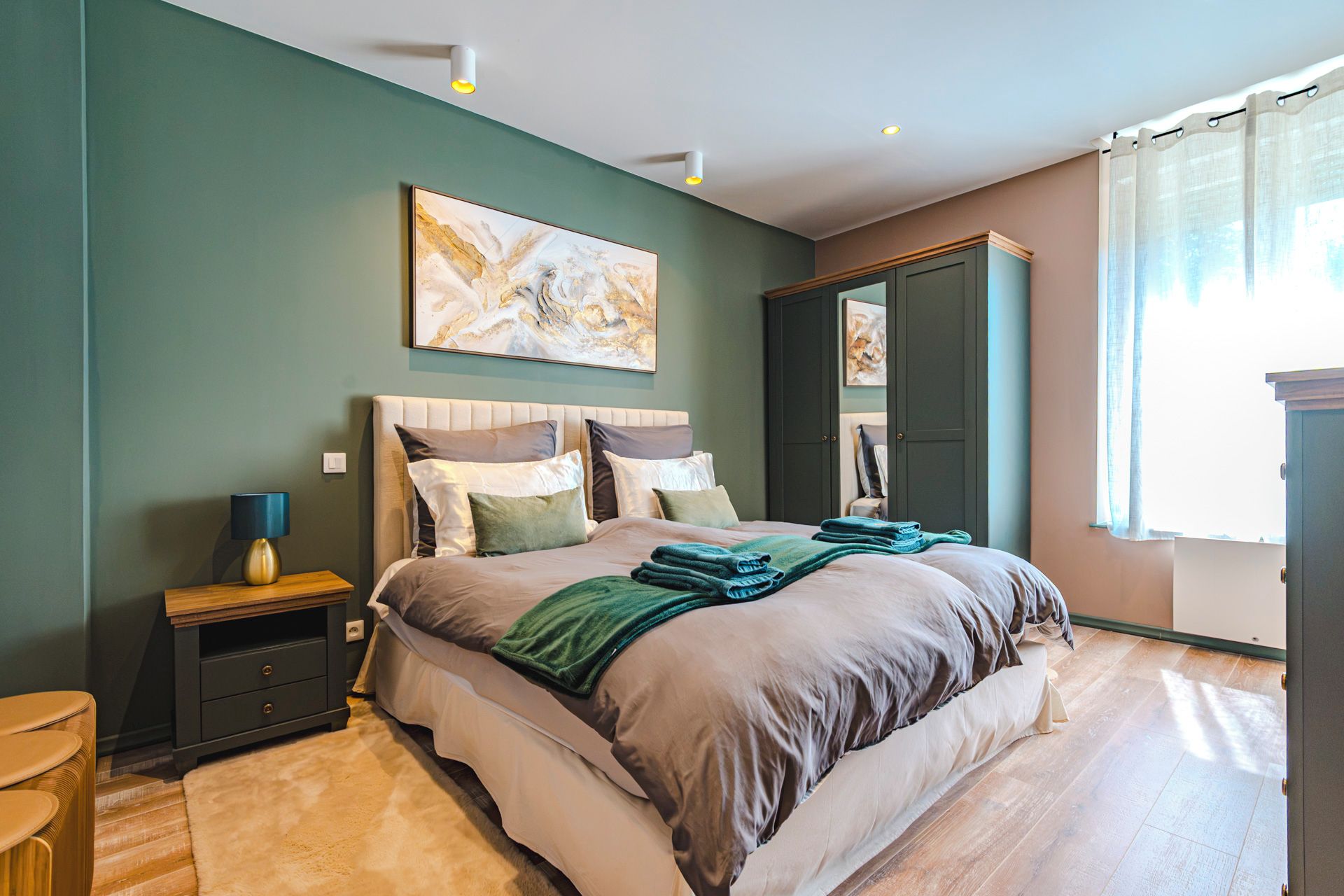 A bedroom with a bed, bedside table, lamp, artwork, wardrobe, and a view of the outdoors.
