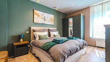 A bedroom with a bed, bedside table, lamp, artwork, wardrobe, and a view of the outdoors.