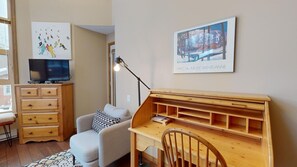 Ein gemütliches Zimmer mit Schreibtisch aus Holz, einem bequemen Sessel, einem Fernseher und Bildern an den Wänden.