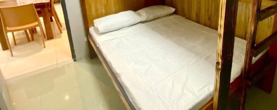 Ein kleines, ordentlich bezogenes Bett mit weißer Matratze und Kissen in einem Zimmer, das ein Holz-Kopfstück hat.