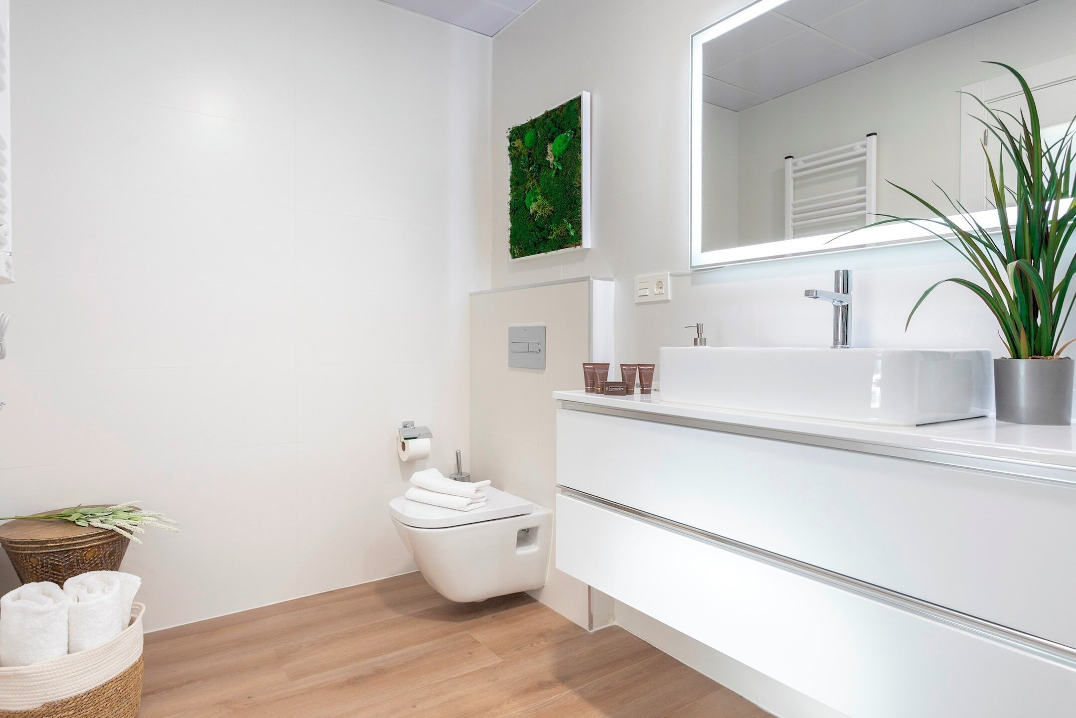 A modern bathroom with a white sink, mirror, and toilet. There is a green wall art piece above the toilet and a potted plant on the sink counter.