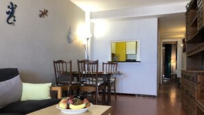A living room with a sofa, a dining table with chairs, a fruit bowl, and a kitchen area with cabinets and a mirror.