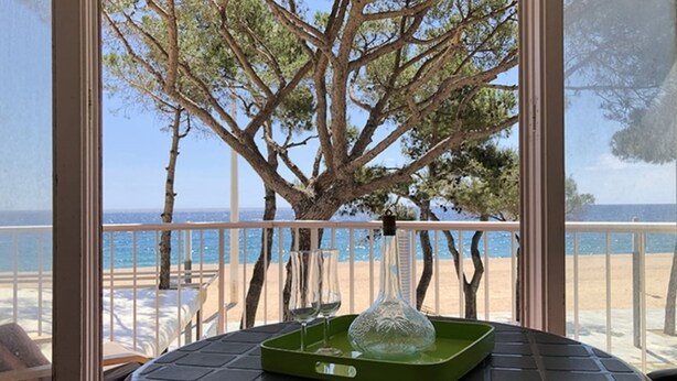 A balcony with a view of the sea, a large tree, and a table with a glass vase and wine glasses.