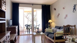 A living room with a sofa, a coffee table, and a television. There is a balcony with chairs and a table outside.