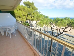 A balcony with a white railing, a small table, and chairs. There are trees and a view of the sea.