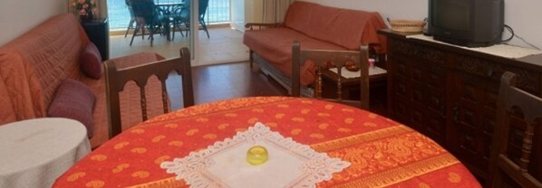 A dining area with a round table covered by a red tablecloth, a white doily, and a yellow candle. There is a wooden chair with a cushion, a sofa, and a television on a cabinet.
