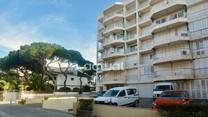 Um edifício residencial com vários andares, varandas, carros estacionados e uma árvore ao fundo.