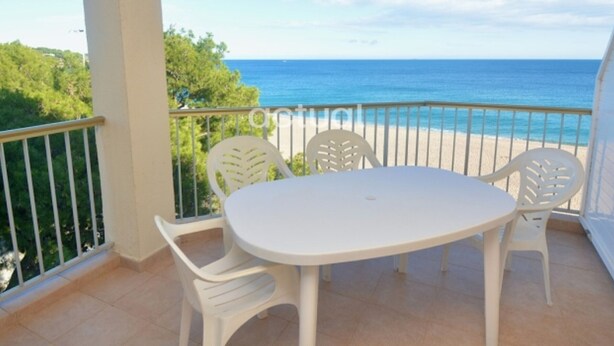Varanda com mesa branca e quatro cadeiras, com vista para uma praia e o mar.