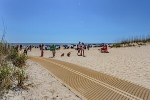 Een zanderig strand met een pad naar het water, parasols en mensen.