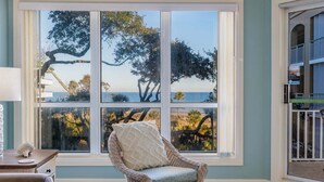 A room with a large bay window overlooking a coastal landscape, a wicker chair with a cushion, and a small table with a decorative item. - Oceanfront Palmetto Dunes Villa w/ Balcony, King Suite & Stunning Views (Hilton Head Island)