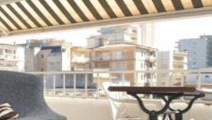 Eine Dachterrasse mit grauem Sofa, einem kleinen Tisch mit bunten Tischdecken und Blick auf Gebäude im Hintergrund.