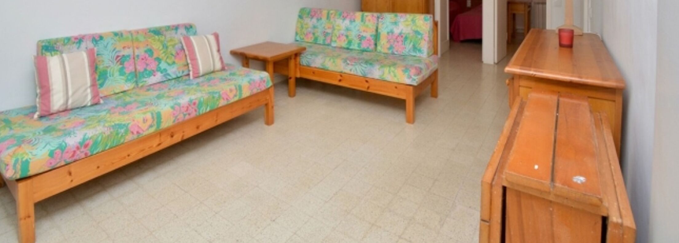 Een kamer met twee houten banken met bloemrijk gestoffeerde bekleding, een klein houten tafeltje en een houten kast.