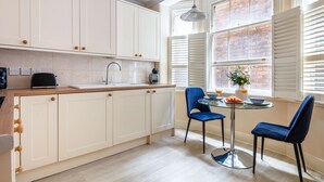 Una cocina con armarios blancos, encimera de madera, un fregadero y una ventana con persianas. Hay una mesa con dos sillas azules y un florero con flores.