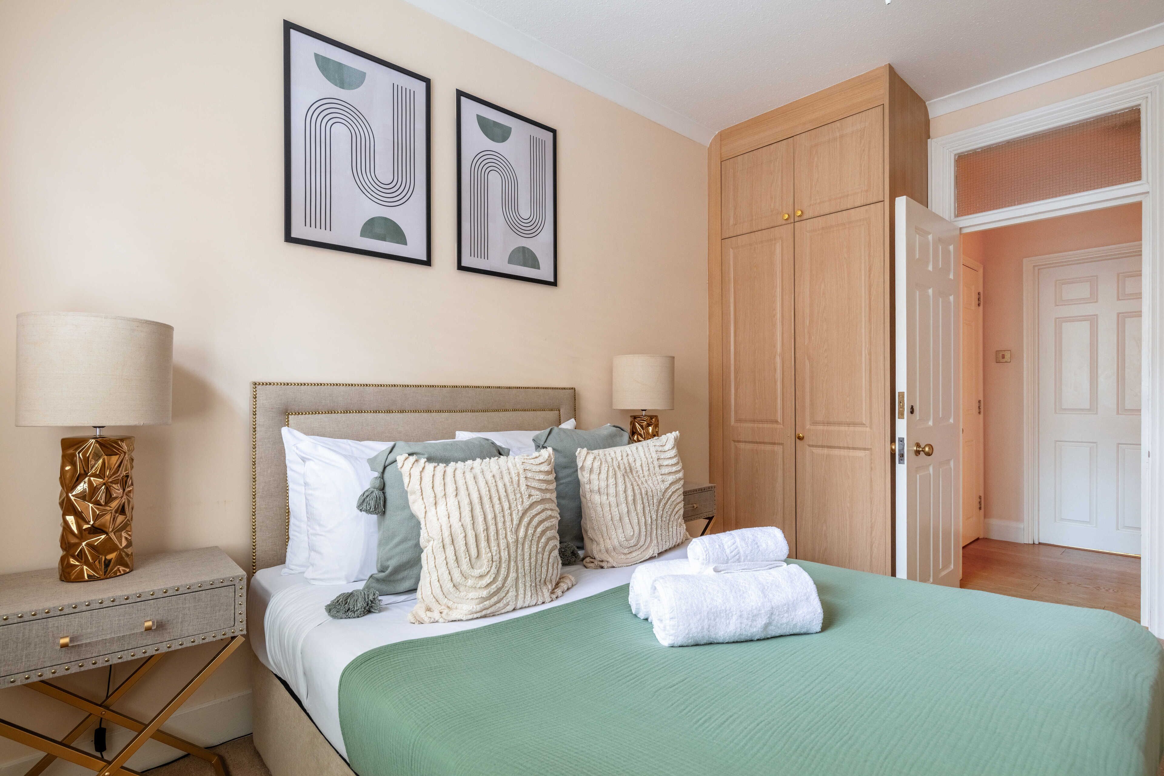 A hotel room with a bed, two bedside tables, a lamp, a green bedspread, and two framed artworks on the wall.