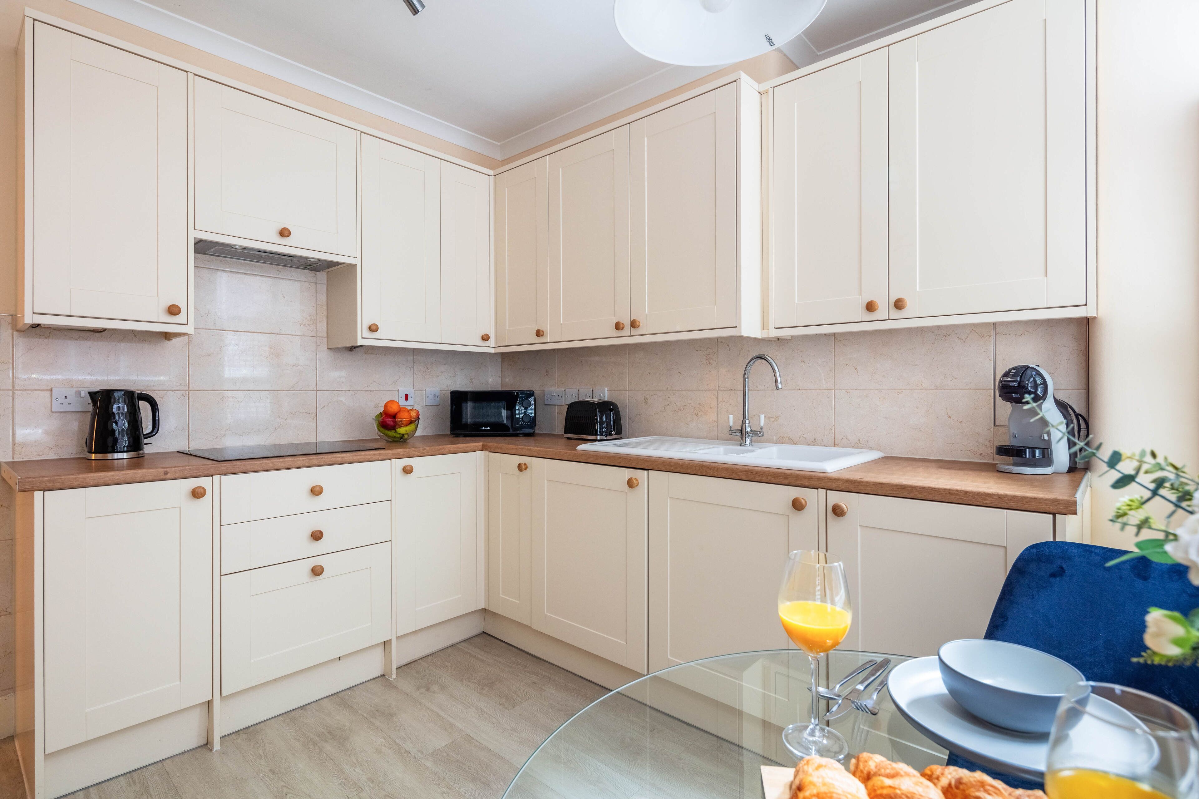 Een moderne keuken met lichte kastjes, een houten aanrecht, een spoelbak en een glazen tafel met ontbijtartikelen.