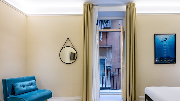 Une chambre d’hôtel avec un canapé bleu, un lit, une grande fenêtre avec des rideaux, une affiche encadrée et un miroir fixé au mur.
