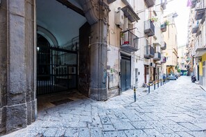 A narrow cobblestone street with a building entrance featuring a wrought iron gate and a small balcony with potted plants.