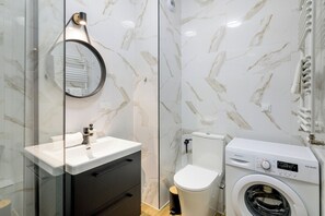 Une salle de bain moderne avec un pare-baignoire en marbre, un miroir rond, un lavabo blanc, un meuble noir, des toilettes et un lave-linge à chargement frontal.