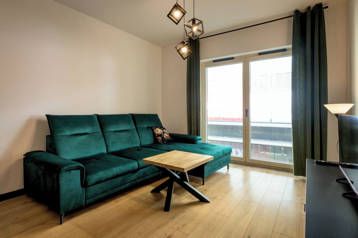 A modern living room with a green sofa, a wooden coffee table, and a large window with curtains.