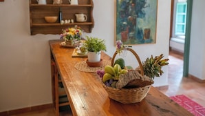 A wooden dining table with a wicker basket containing fruits and a potted plant. A wooden shelf with kitchenware and a painting is in the background. - Apartment in Tuscany Near Pisa and Beaches (Collesalvetti)