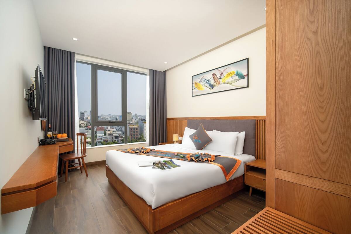 Ein Hotelzimmer mit einem großen Bett, einem Schreibtisch, einem Stuhl, einem Fernseher und einem Fenster mit Blick auf die Stadt.