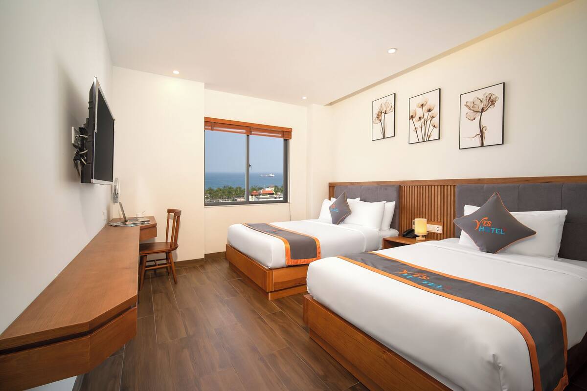 Ein Hotelzimmer mit zwei Betten, einem Schreibtisch, einem Sessel und einem Fernseher. An den Wänden hängen gerahmte Bilder und das Fenster bietet einen Ausblick.