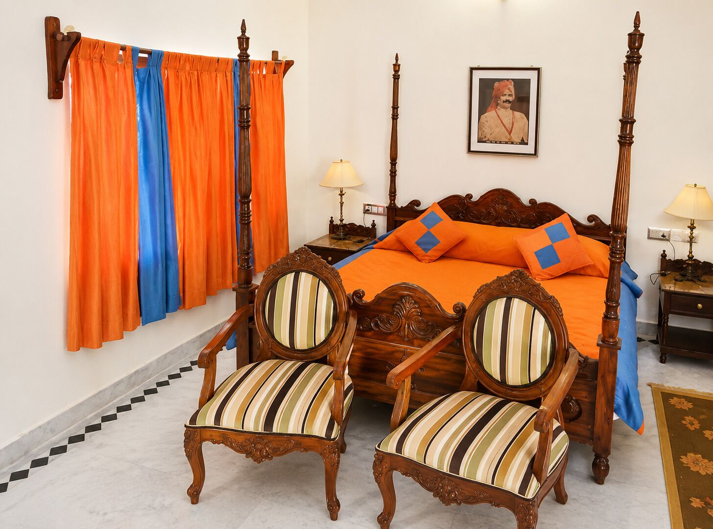 Uma cama de dossel com cabeceira de madeira, duas poltronas estofadas com almofadas listradas, um quadro na parede e um abajur sobre uma mesa de cabeceira.