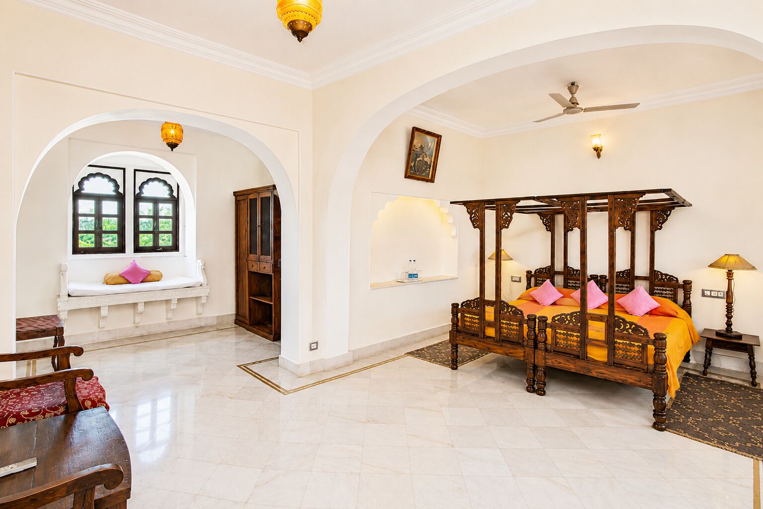 A spacious room with a large bed, a seating area with a bench, and a view through an arched window.
