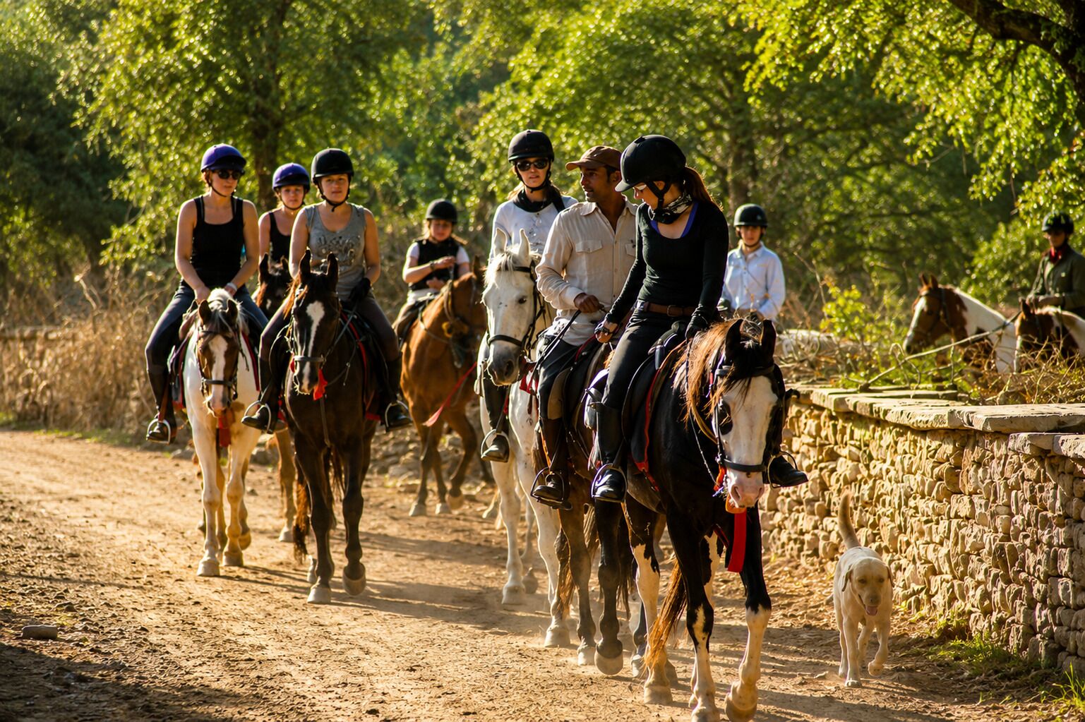 A group of people riding horses on a dirt path with a stone wall on one side.