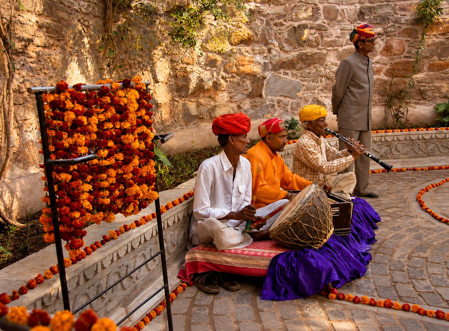 Un muro de piedra con una barandilla metálica decorativa, un grupo de personas tocando instrumentos musicales tradicionales, y un hombre tocando una flauta.