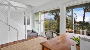 Un intérieur moderne avec un escalier, un coin repas comprenant une table et des chaises, et une grande porte-fenêtre coulissante donnant sur un balcon avec vue sur les arbres et les collines.