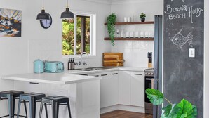 Une cuisine moderne avec des meubles blancs, un grand îlot central, des tabourets de bar, un tableau noir sur lequel on peut lire « Bienvenue à la maison de plage de Boronia », et une fenêtre donnant sur un espace verdoyant.