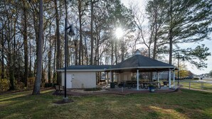 Ein Gazebo mit überdachtem Picknickbereich, ein kleines Gebäude und eine von Bäumen umgebene Rasenfläche.
