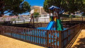 Ein eingezäunter Spielplatz mit Rutsche und Klettergerüst, umgeben von Bäumen und Gebäuden.