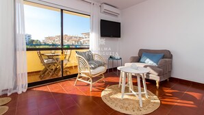 Ein Balkon mit Blick auf Gebäude, einer Couch, einem Sessel, einem kleinen Tisch und einem an der Wand montierten Fernseher.