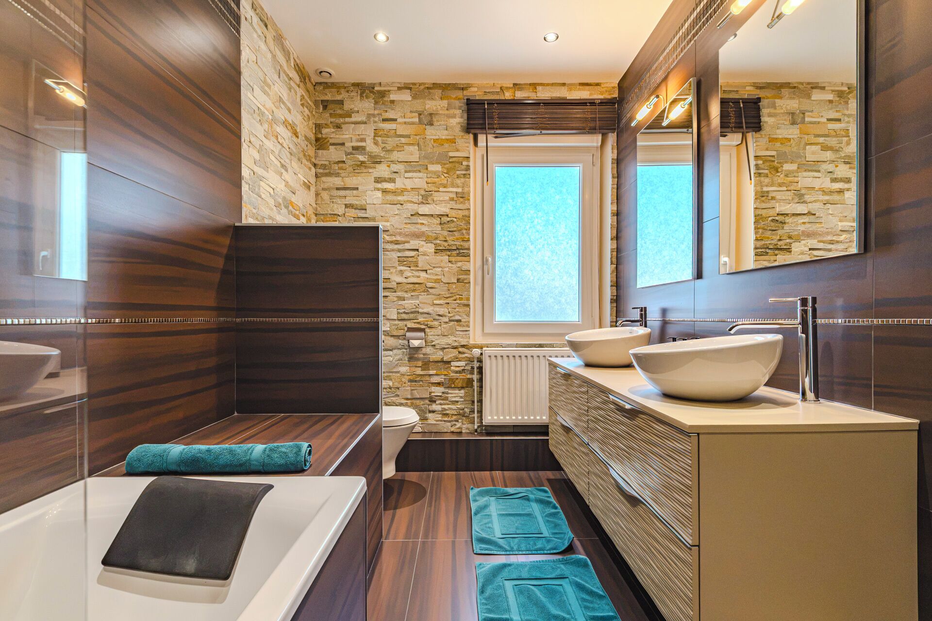Een moderne badkamer met een stenen muur, een grote spiegel, een bad, twee wastafels en een raam.