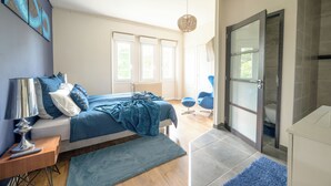 Une chambre moderne avec un grand lit, une table de chevet avec une lampe, et une vue sur la salle de bain à travers une porte ouverte.
