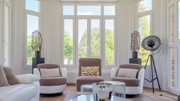 Un salon moderne avec deux canapés blancs, un fauteuil marron et une table basse en verre. De grandes fenêtres offrent une vue sur les arbres et un mur en briques.