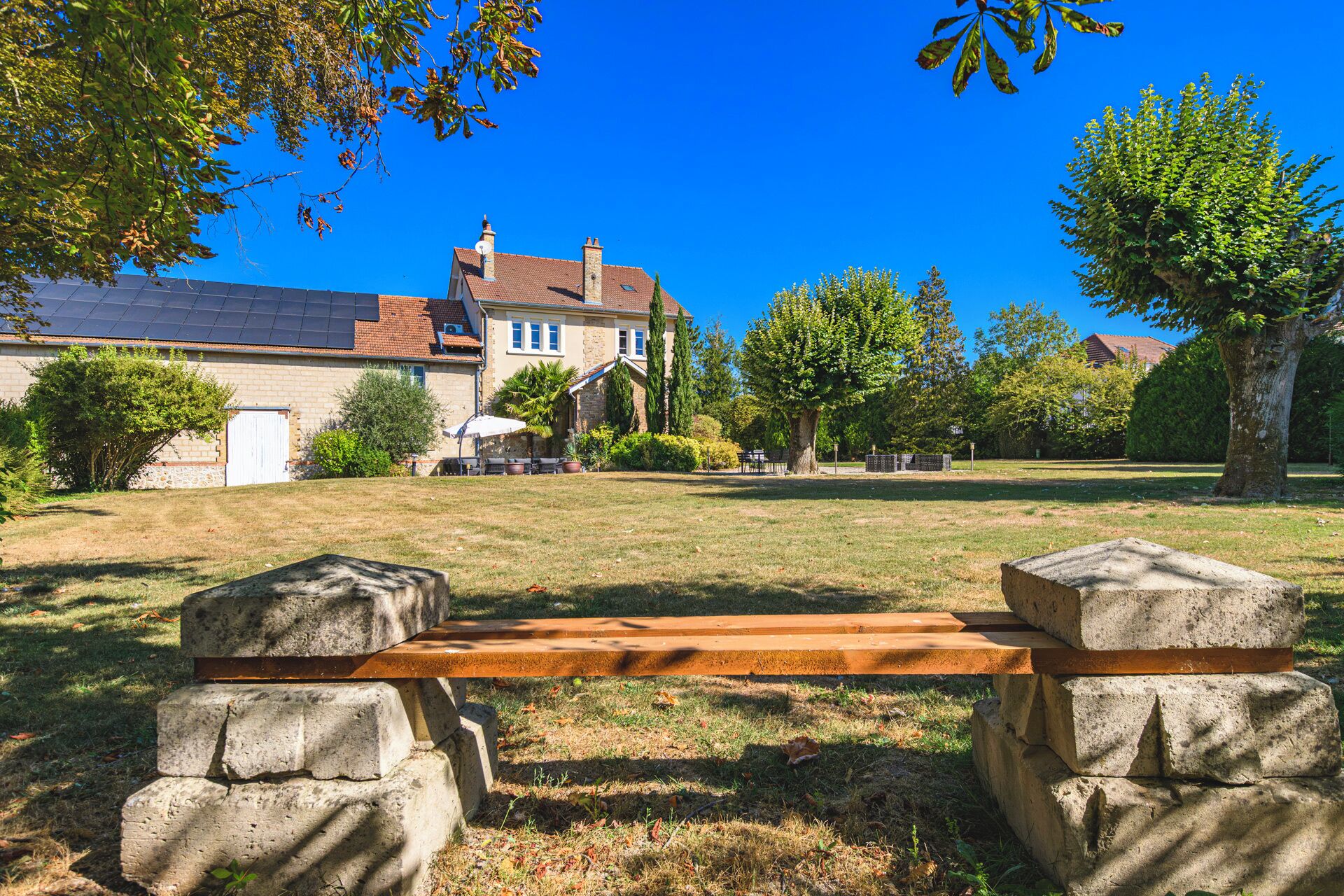 Un banc avec des supports en pierre, situé dans un espace verdoyant, avec une maison en arrière-plan.