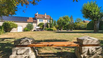 Un banc avec des supports en pierre, situé dans un espace verdoyant, avec une maison en arrière-plan.