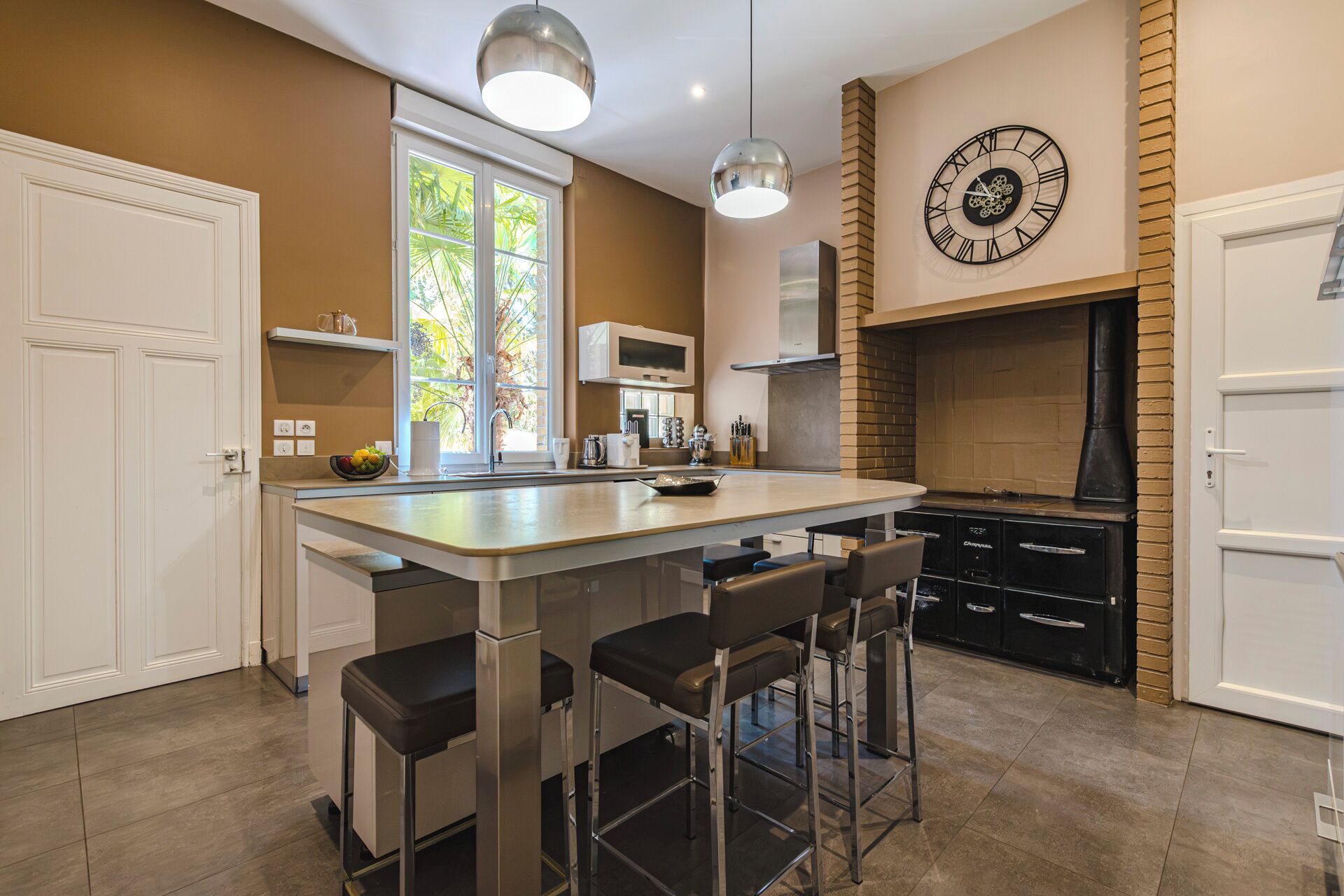 A modern kitchen with a central island, bar stools, a brick fireplace, and a large wall clock.