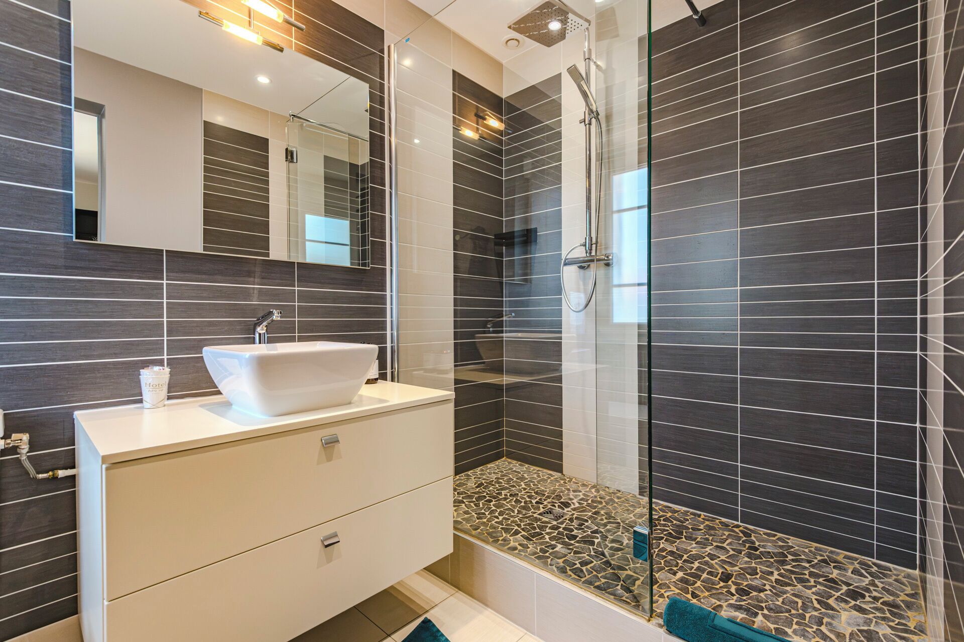 Une salle de bain moderne avec un meuble-lavabo blanc, une cabine de douche en verre et des murs recouverts de carreaux foncés.