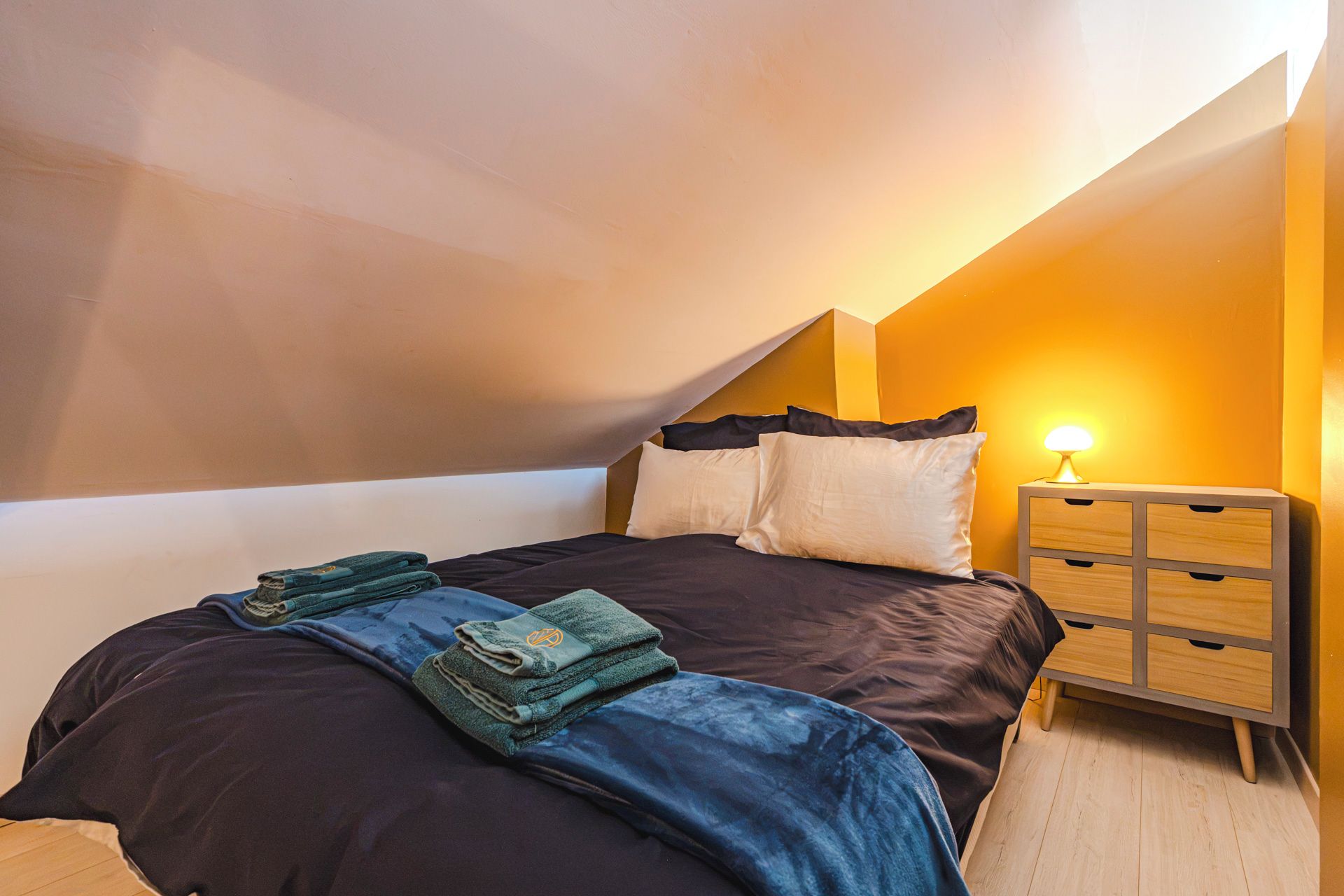 A bedroom with a bed, pillows, a bedside table with a lamp, and a wooden nightstand.