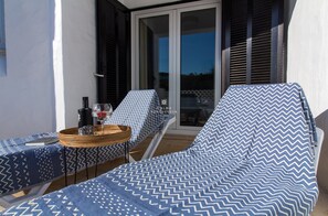 A balcony with two lounge chairs, a table with a bottle and glass, and a window with blinds. - Balaia Golf Village - Blue Apartment (Albufeira)