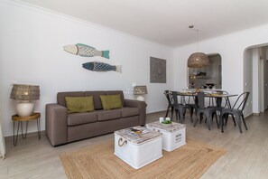 A living room with a sofa, coffee table, dining table, chairs, and decorative wall art. - Balaia Golf Village - Blue Apartment (Albufeira)