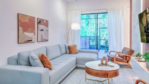 A modern living room with a light grey sectional sofa, a round coffee table, and a brown armchair. There are abstract paintings on the wall and a large window with curtains.