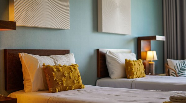 Two beds with white and yellow pillows, a nightstand with a lamp, and a wall with textured artwork.