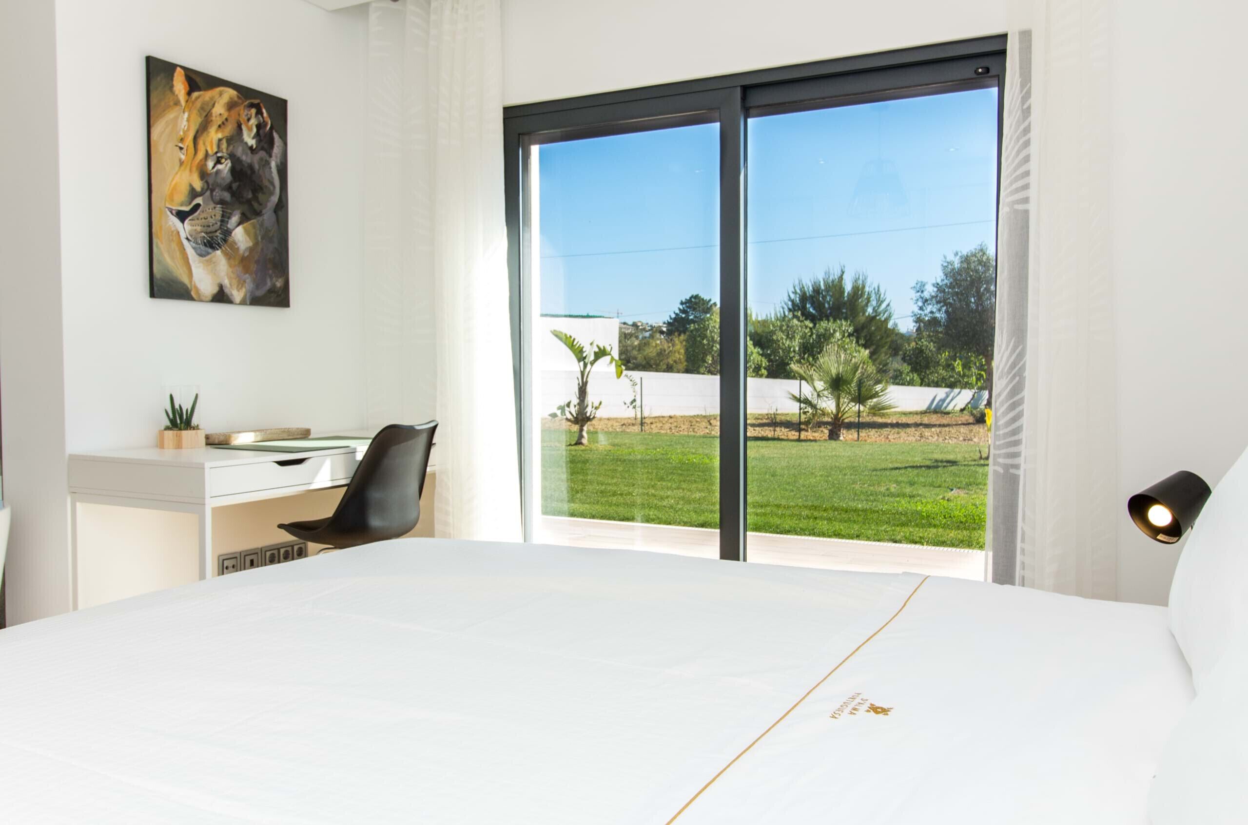 A bedroom with a large bed, a desk, a chair, a painting, and a view of a green landscape through a sliding door.
